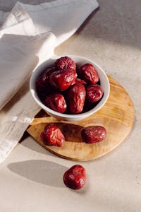 A ceramic bowl filled with red dates on a wooden board, casting soft shadows.